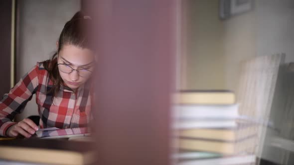 A girl sits at a table with books and studies information on a tablet screen