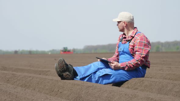 Farmer, Agronomist Sits Between Special Soil Rows on Field. He Tests, Using Tablet, Quality