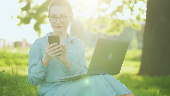 Busy Attractive Woman Working at the Laptop and Using Smartphone While Sitting on Grass in City Park