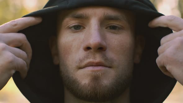 Young Bearded Guy with Freckles Putting on Black Hood and Looking Seriously to Camera Standing