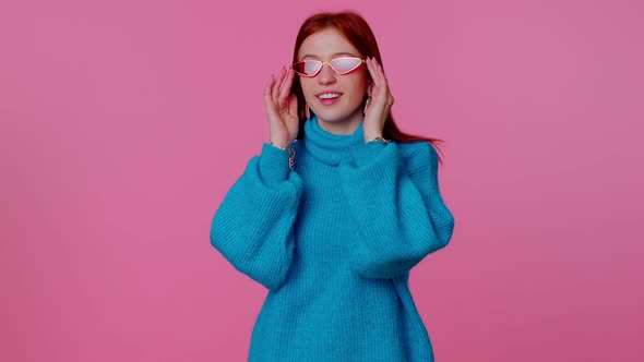 Seductive Cheerful Redhead Girl in Blue Sweater Wearing Sunglasses Charming Smile on Pink Wall