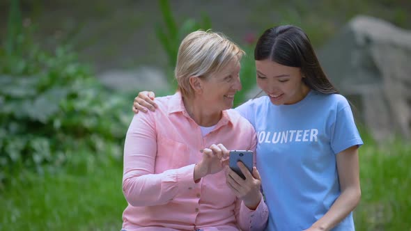 Mature Woman and Young Volunteer Looking Smartphone Photos Together, Assistance