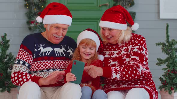Smiling Grandparents and Granddaughter Waving During Video Call on Smartphone at Christmas Reunion