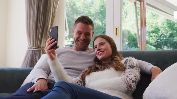 Caucasian couple smiling while having a video call on smartphone sitting on the couch at home