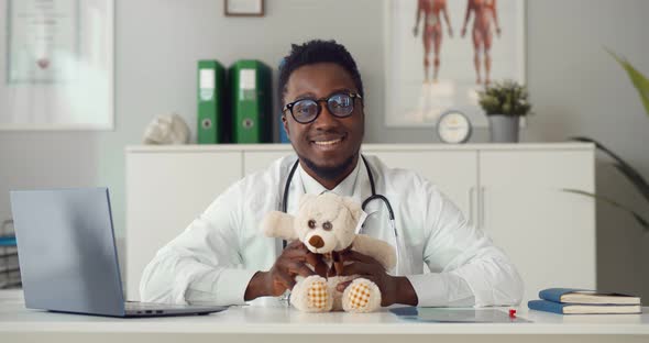 Portrait of African Young Pediatrician Sitting at Desk with Teddy Bear Smiling at Camera