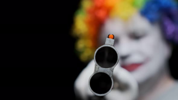 smiling evil clown holds gun aiming barrel at camera 