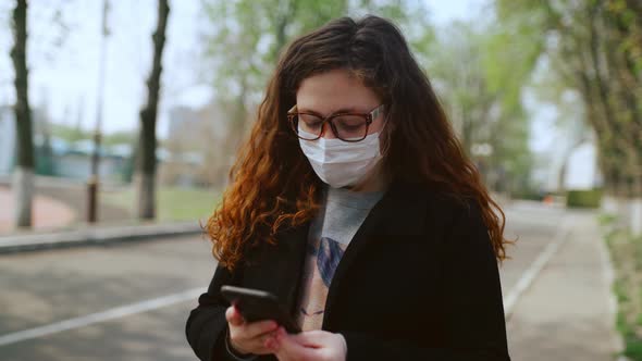 Young Girl in a Medical Mask Uses a Smartphone in the Park