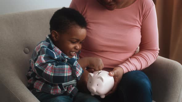 Little Black Boy Putting Coins Into Piggy Bank While Sitting with Mom