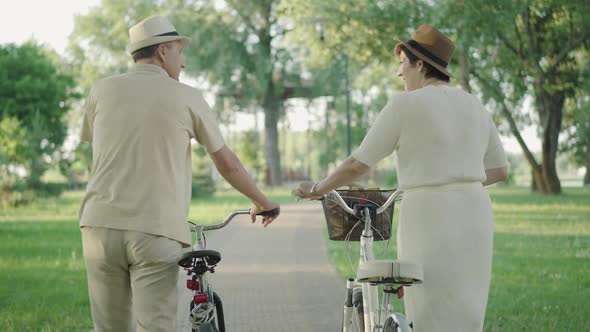 Back View of Relaxed Confident Mid-adult Couple Walking with Bikes Along the Park Alley. Camera