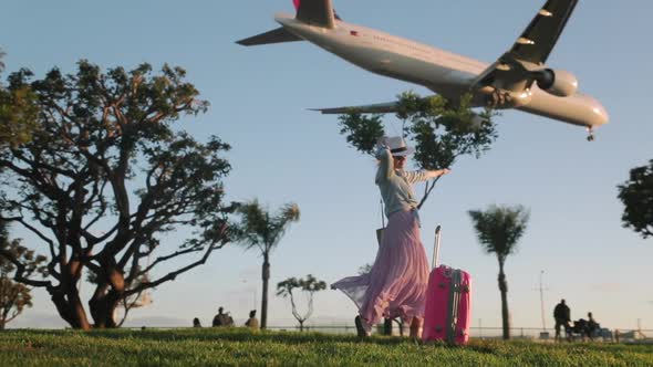 Beautiful Woman with Pink Travel Bag Is Happy To See Landing Airplane at Sunset