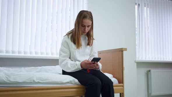 Absorbed Young Woman Messaging Online on Smartphone Sitting in Hospital Ward Indoors