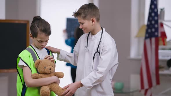 Sad Caucasian Boy Caressing Teddy Bear As Positive Friend in Doctor Gown Supporting Talking