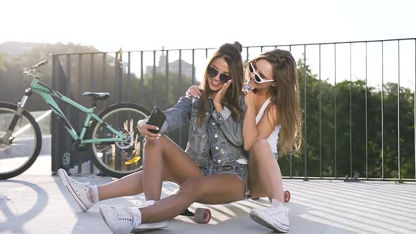 Hot Girlfriends in Trendy Sunglasses and Stylish Clothes Sitting on Skateboard