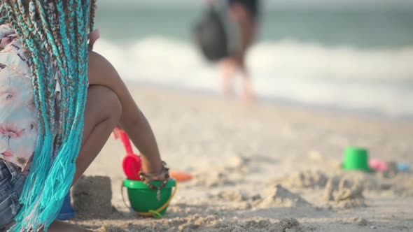 A Girl with African Braids in a Summer Costume Plays on the Beach with Shells Near the Sea with