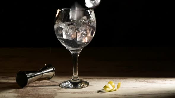 Person pouring ice cubes into glass of gin tonic with lemon and jigger on table in dark room