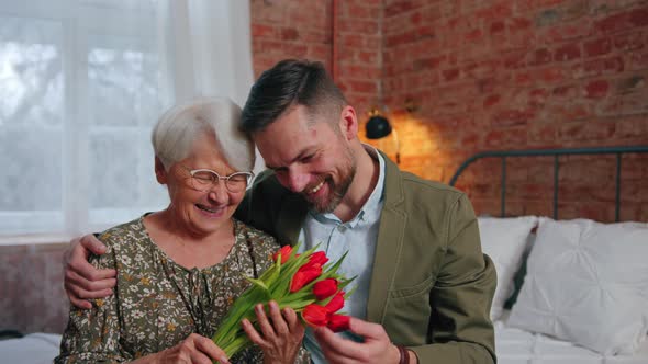 Elderly Pensioner Lady and Her Millennial Son Sitting on a Bed Hugging and Celebrating Mother's Day