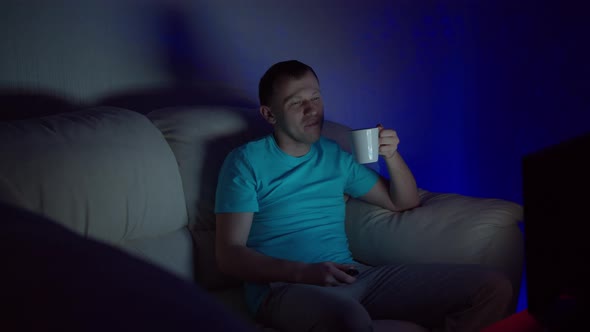 A man with a mug of hot tea sits at the TV late in the evening