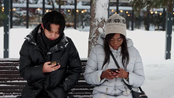 Young Asian Man and Young Black Woman Sitting on Bench in Winter Park and Looking at Their Phones