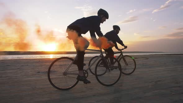 Two Young Men Riding Bicycles on the Beach with Orange Smoke During Sunset