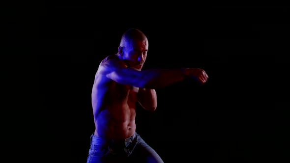 a Muscular Bald Man with a Bare Torso and Jeans Strikes a Combination of Blows on a Black Background