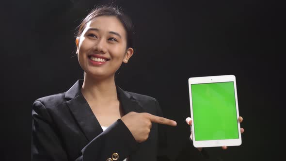 Asian Speaker Woman In Business Suit Holding And Pointing Green Screen Tablet In The Black Studio