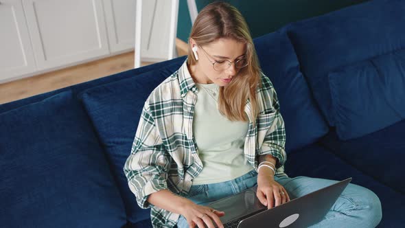 Top View of Young Focused Lady Wearing Earphones Typing on Laptop Computer Sitting on Sofa at Home