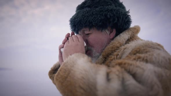 Side View Closeup Bearded Old Man Drinking Hot Tea with Cloudy Winter Sky and Ice at Background