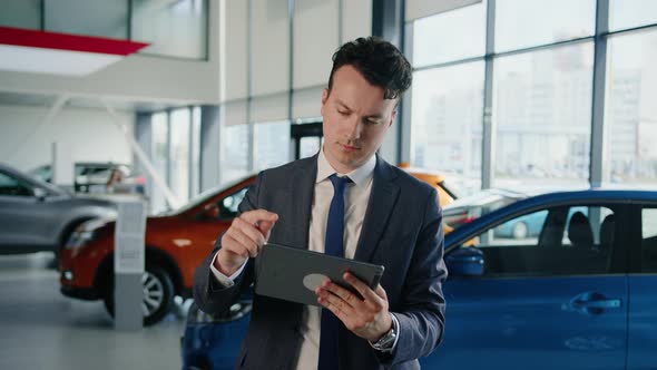 General Manager in a Suit and Tie with a Tablet in His Hands in a Car Dealership