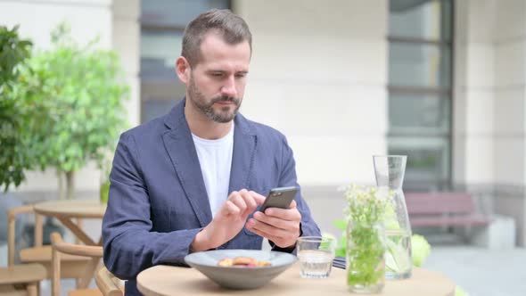 Man Browsing Internet on Smartphone Sitting in Outdoor Cafe Front Pose