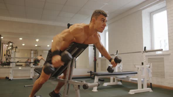 Young Muscular Man with a Naked Torso Works Out in the Gym and Does Arm Lift with Dumbbells Lying on