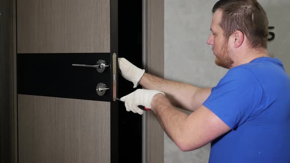 A Carpenter In A Special Uniform Repairs The Lock Of The Front Door. Repair Of The Door Lock