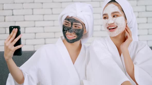 Two happy Asian girls in white bathrobes with facial mask taking photo on smartphone together