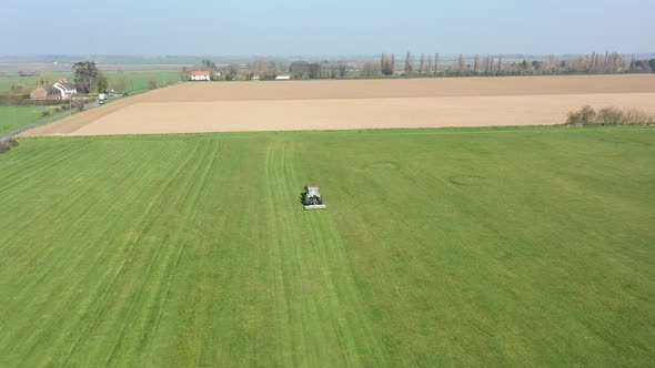 Tractor mowing green field drone hovering static. Trees in distance, rural village HD60