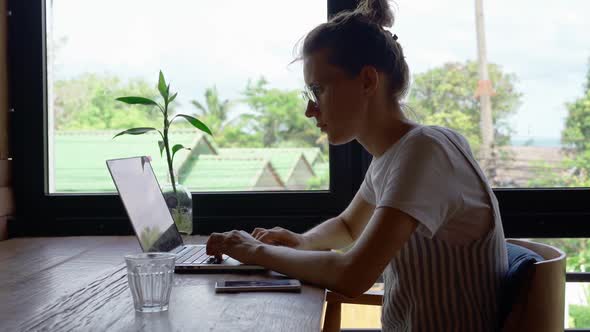 Female freelancer sitting at table near window and browsing data on laptop in cafeteria