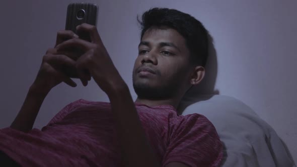 Young Indian teenager laying back on the bed and using mobile while sleeping during the night on bed