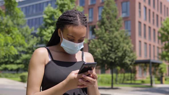 A Young Black Woman in a Face Mask Takes Selfies with a Smartphone - an Office Building and Trees