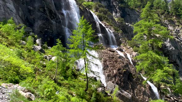 Tilt up shot of green fir trees and falling waterfall down high mountains in Austria during sunny da