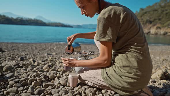 Woman Hiker Tourist Pours Coffee From Coffee Maker Into Cups on Shores of Mediterranean Sea on