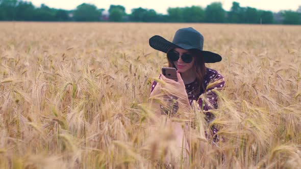 Beautiful Young Hippie Woman in the Wheat Field at Sunset Uses a Smartphone