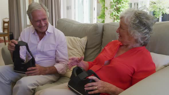 Happy caucasian senior couple sitting on couch in living room holding vr headsets and talking