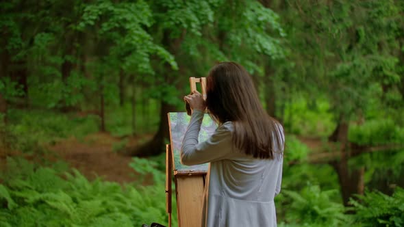 Rear View of a Young Artist Woman Painting a Landscape in a Summer Forest