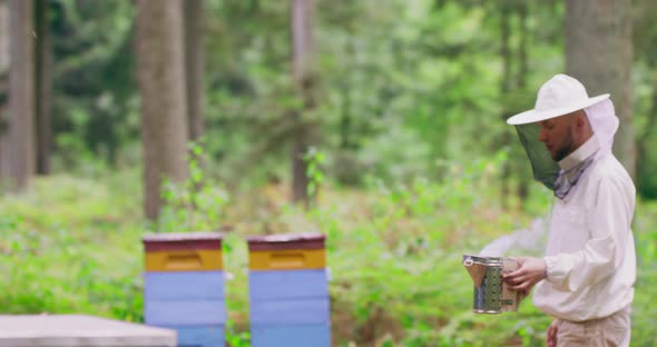 Bearded Young Male Beekeeper in White Protective Suit Walks Through the Apiary Between the Hives in