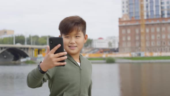 Asian Boy Walking along Riverside and Video Calling on Smartphone