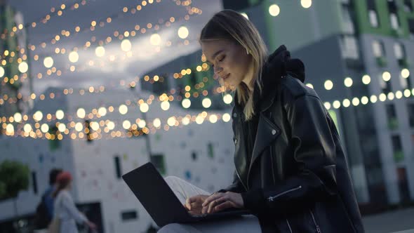 Young Girl in Wireless Headphones Uses a Laptop Sitting Outside in a Modern Residential Area Evening