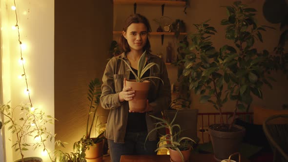 A Happy Young Girl Stands in the Middle Her Home Greenhouse and Holding a Clay Pot with a Houseplant