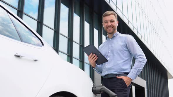 Young European Businessman with Wireless Headphones and Tablet Near Electric Car Charging the