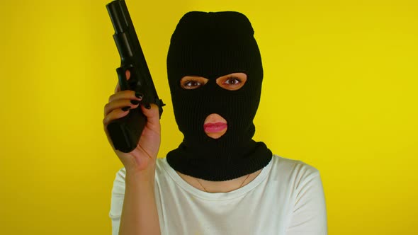 Unrecognizable woman in black balaclava with gun on yellow background.