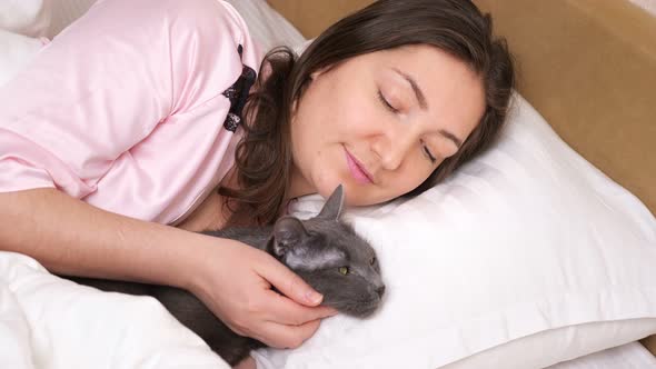 Relaxed Woman in Pale Pink Pajama Pets Sleepy Cat in Bed