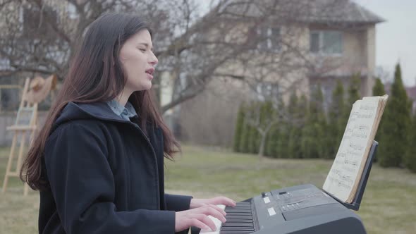 A Successful Brunette Girl Plays on a Synthesizer and Sings While Sitting in the Backyard