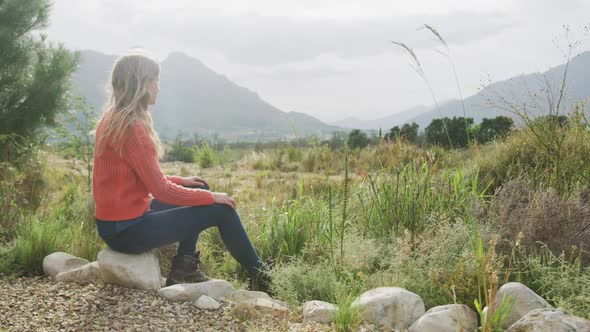 Caucasian woman having a good time on a trip to the mountains, sitting on a rock, admiring the view